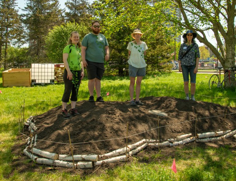 A row of four people stand at a garden plot at the Goldenrod Community Garden. It is a sunny day and there are trees behind them.