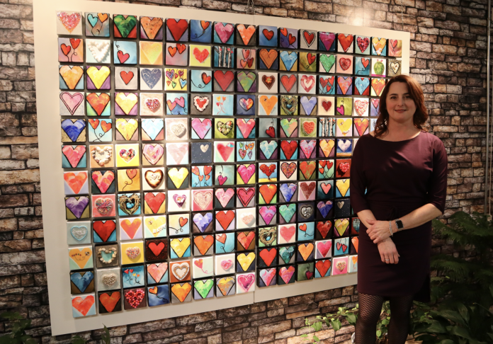 Alison stands next to a wall full of her hearts. It’s very colourful.