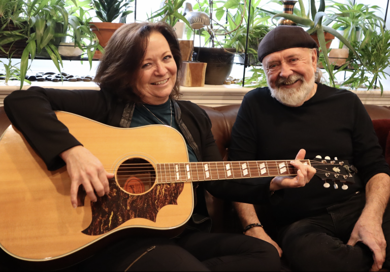 Sherri and dick pose for a photograph on a couch. Their are plans in the background. She’s holding a guitar.