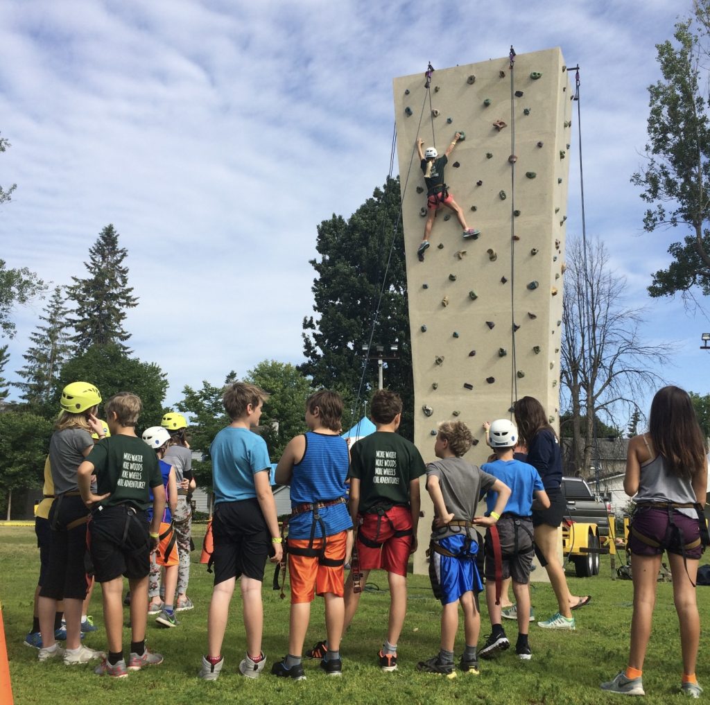 A child climbs up a rock wall as many other kids look on.