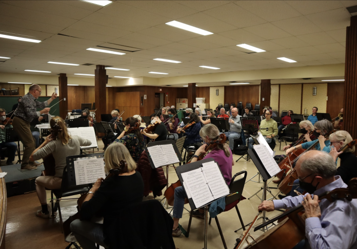 A group of orchestra members perform together|A man in a flashy gray shirt conducts a community orchestra||A man wearing his burgundy baseball cap backwards plays the oboe|||Two men in face coverings play the violin.|A man in a gray shirt plays the bass clarinet.