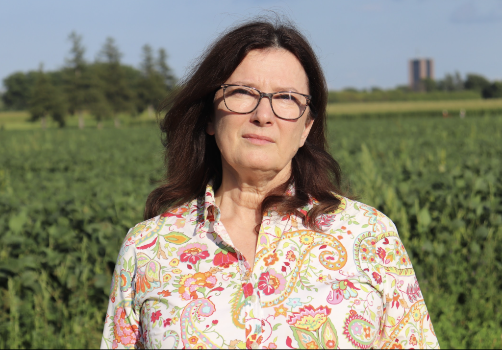 Karen who is wearing a very colourful and flashy shirt poses for a photograph with the experimental farm in the background. Dunton Tower at Carleton University can be seen in the distance.