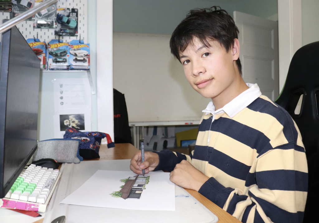 Kai sits at his desk and sketches a house. He’s looking at the camera. 