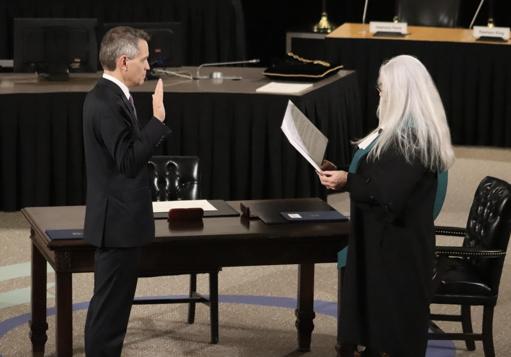 Mark Sutcliffe raises his right hand as he’s sworn into office. A woman with long white hair is wearing a robe.