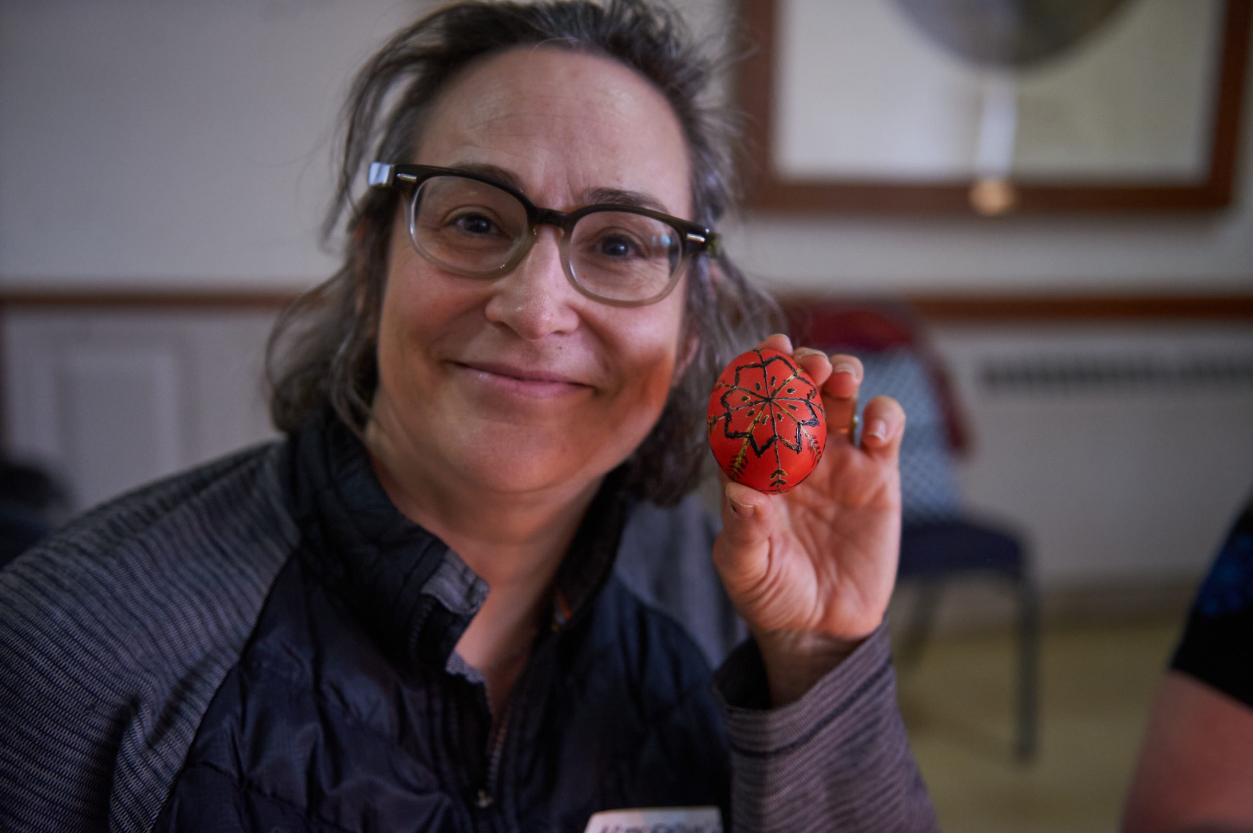 A grey haired women in glasses hoods up a red painted Easter egg with designs on it.