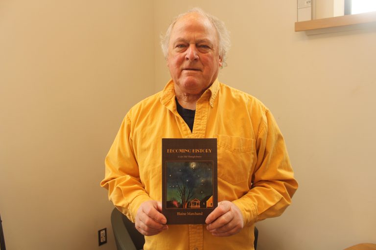 Blaine poses for a photo holding his book. He’s standing next to a wall and is wearing a bright yellow shirt.