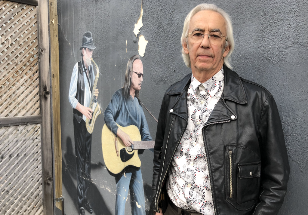 A man in a black leather jacket and a flashy shirt poses for a photograph next to a wall with a mural. It has figures of people playing music painted on it.