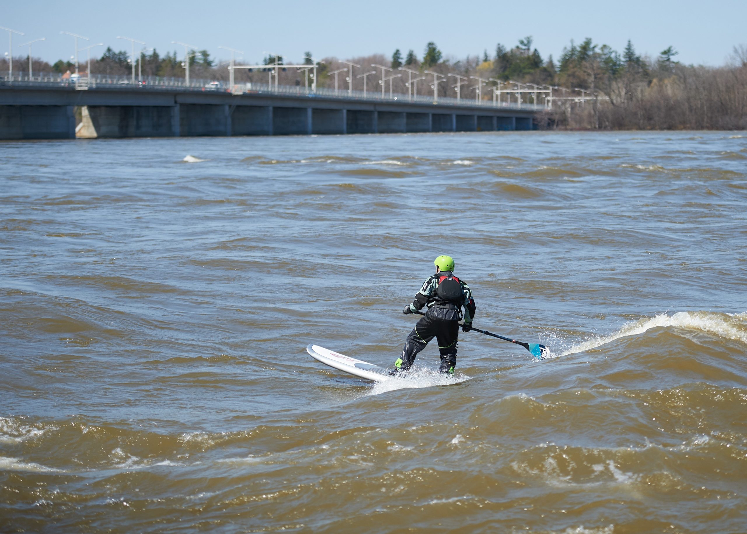 A man paddle boarding at Remic Rapids. The bridge is in the distance to the top left. 