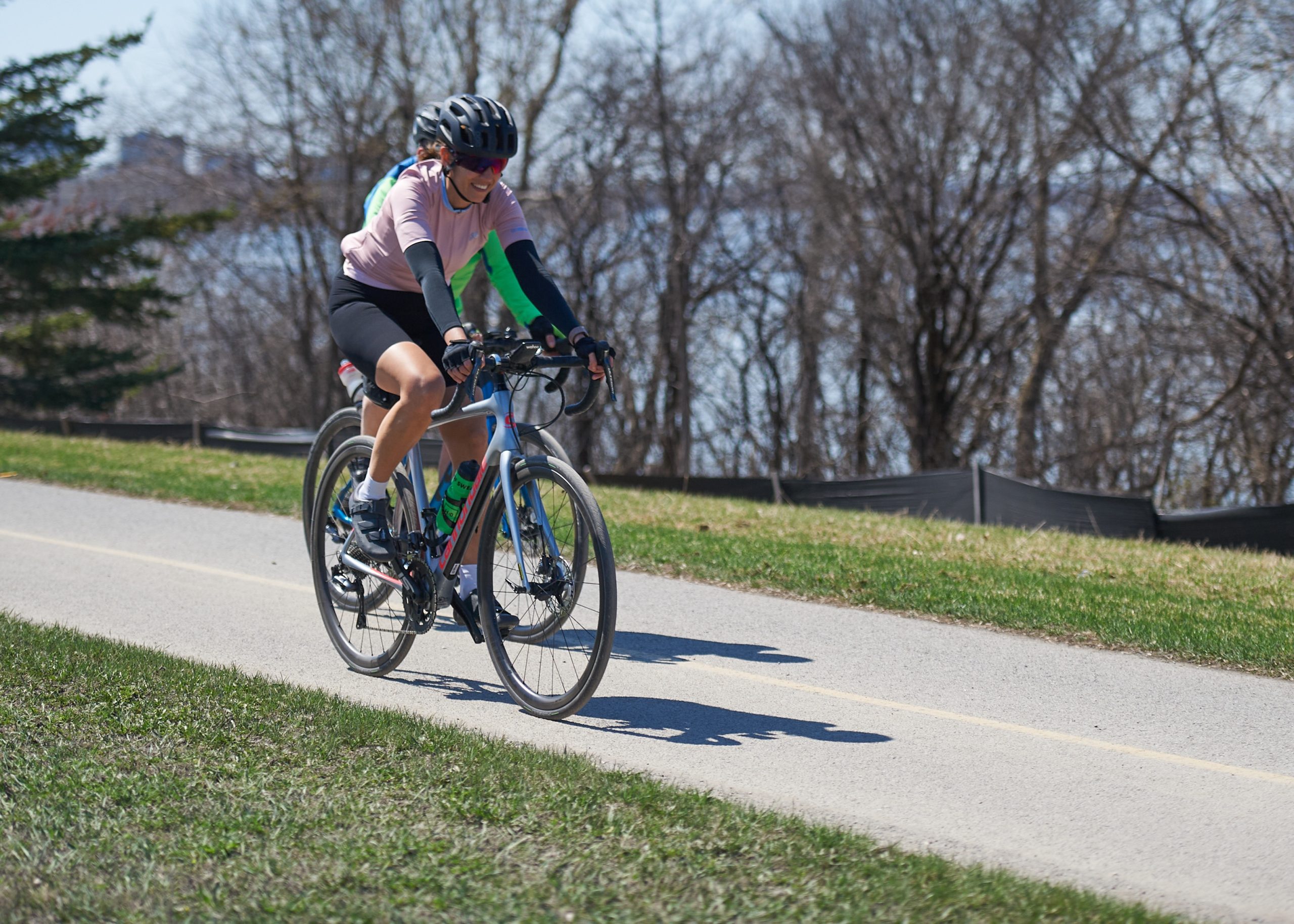 A woman wearing a pink shirt smiles as she bikes down a path. A person can barely be seen behind her, also on a bicycle. 