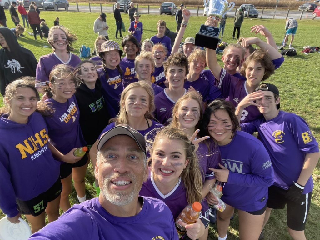 A man takes a selfie with some of his students. They are wearing purple shorts and standing on a field.