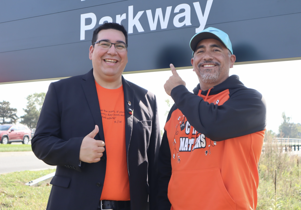 Dylan Whiteduck smiles as he poses for a photograph with another man. Both are wearing orange and giving the thumbs up. 