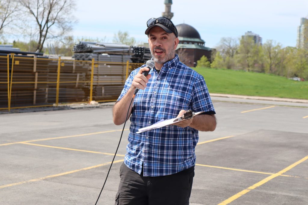 Dave Allston, wearing a blue plaid shirt, stands in a parking lot and talks to the attendees who are out of view in the picture. A mosque can vaguely be seen in the background.