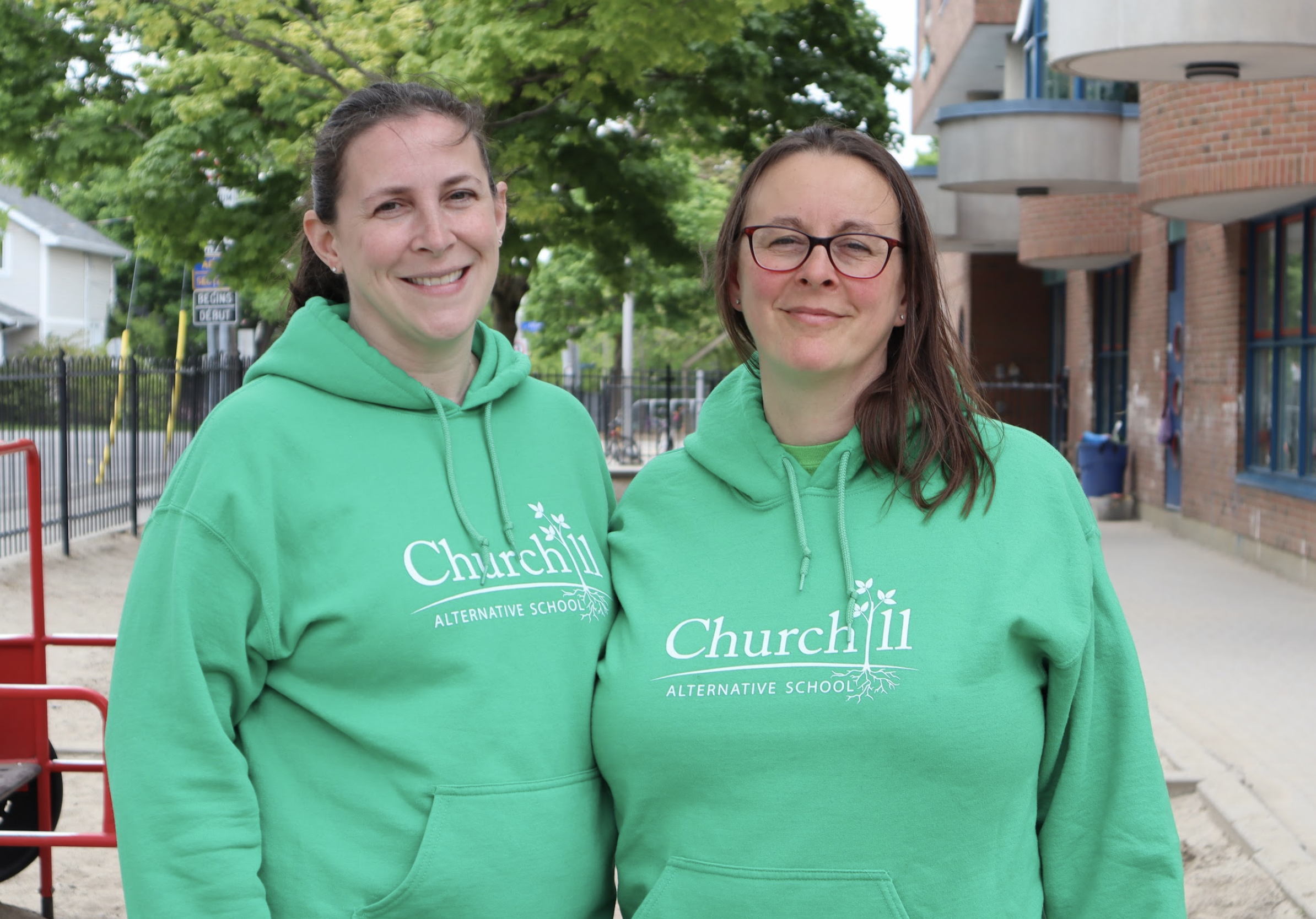 Amanda and Tami pose for a photograph in the school yard. Both are wearing green hoodies with the school’s name on them.