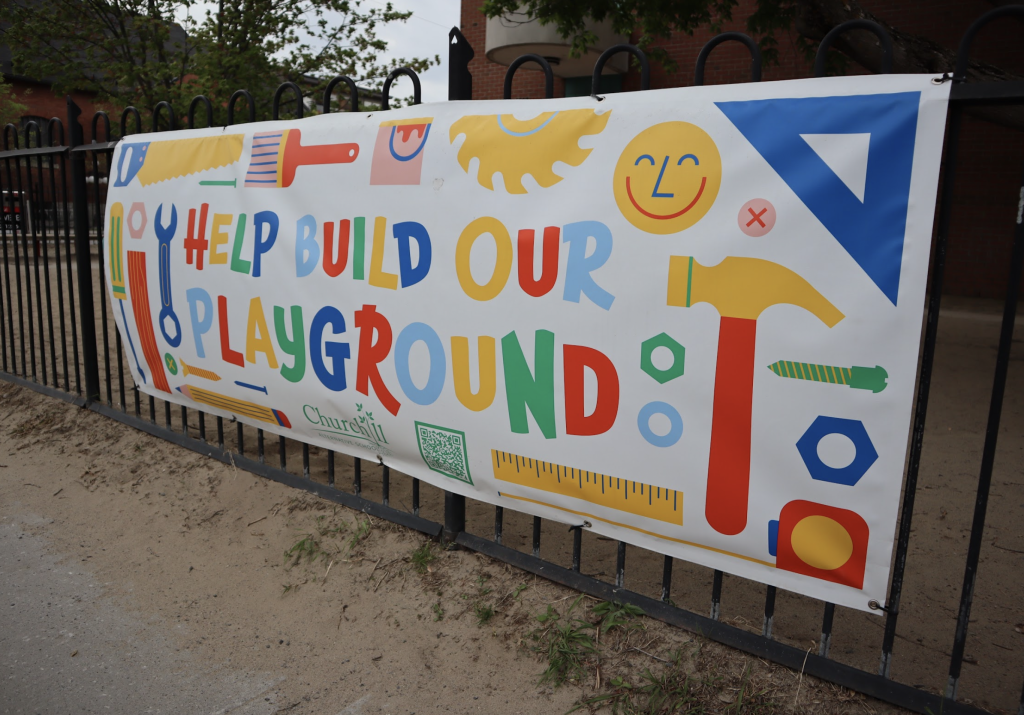 A banner hangs on the fence of the school yard reading “Help build our playground.”