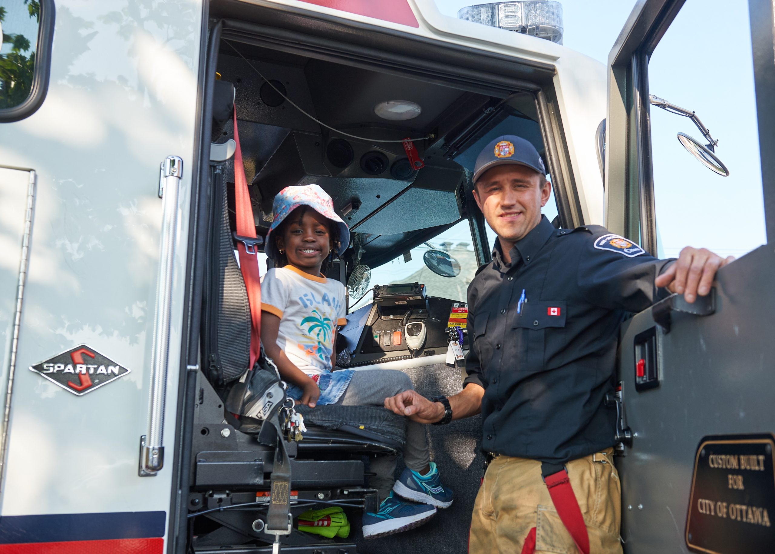 A kid and a firefighter pose for a photograph. The kid is sitting in the fire truck.