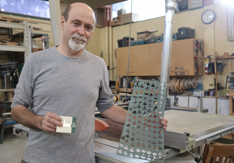 John poses for a photograph in his workshop. He’s holding a green copper sheet with maple leafs pinched out of it.