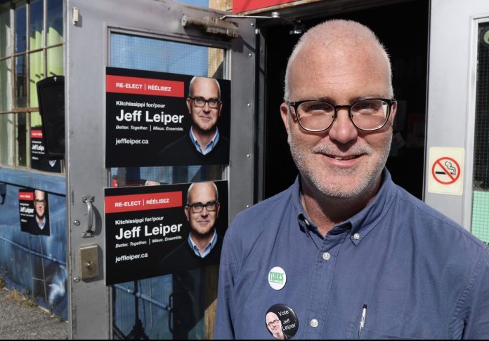 A man running for city council wears a blue shirt and stands next to campaign signs taped to a door.