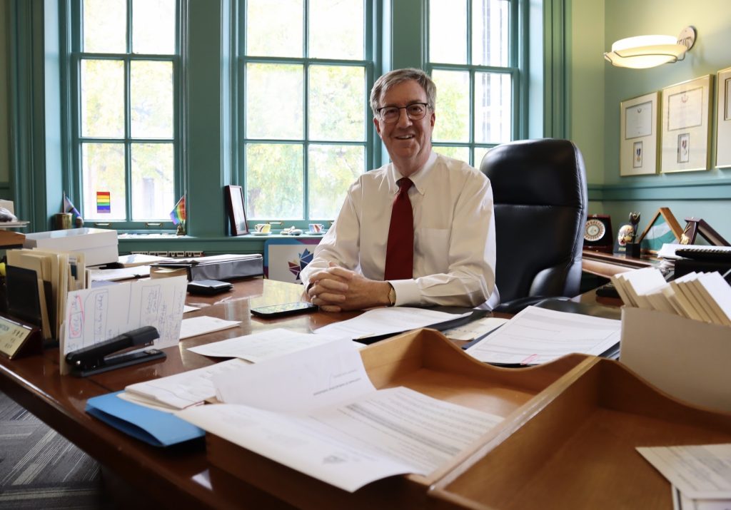 A man wearing a white shirt and read tie poses for a photograph at his desk. Many papers are on the table. 