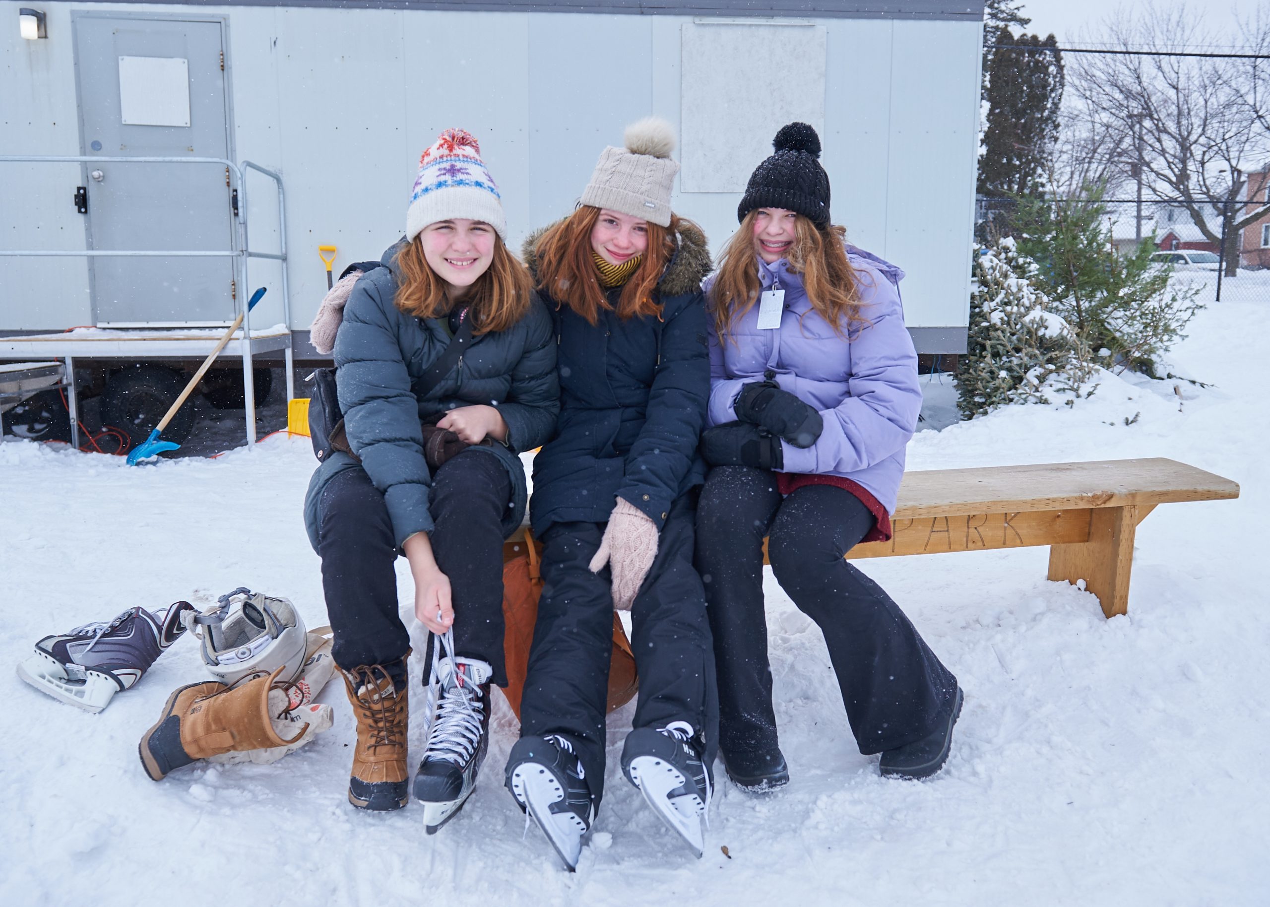 Three girls pose for a photograph as they sit down on a wooden bench to lace up their skates.