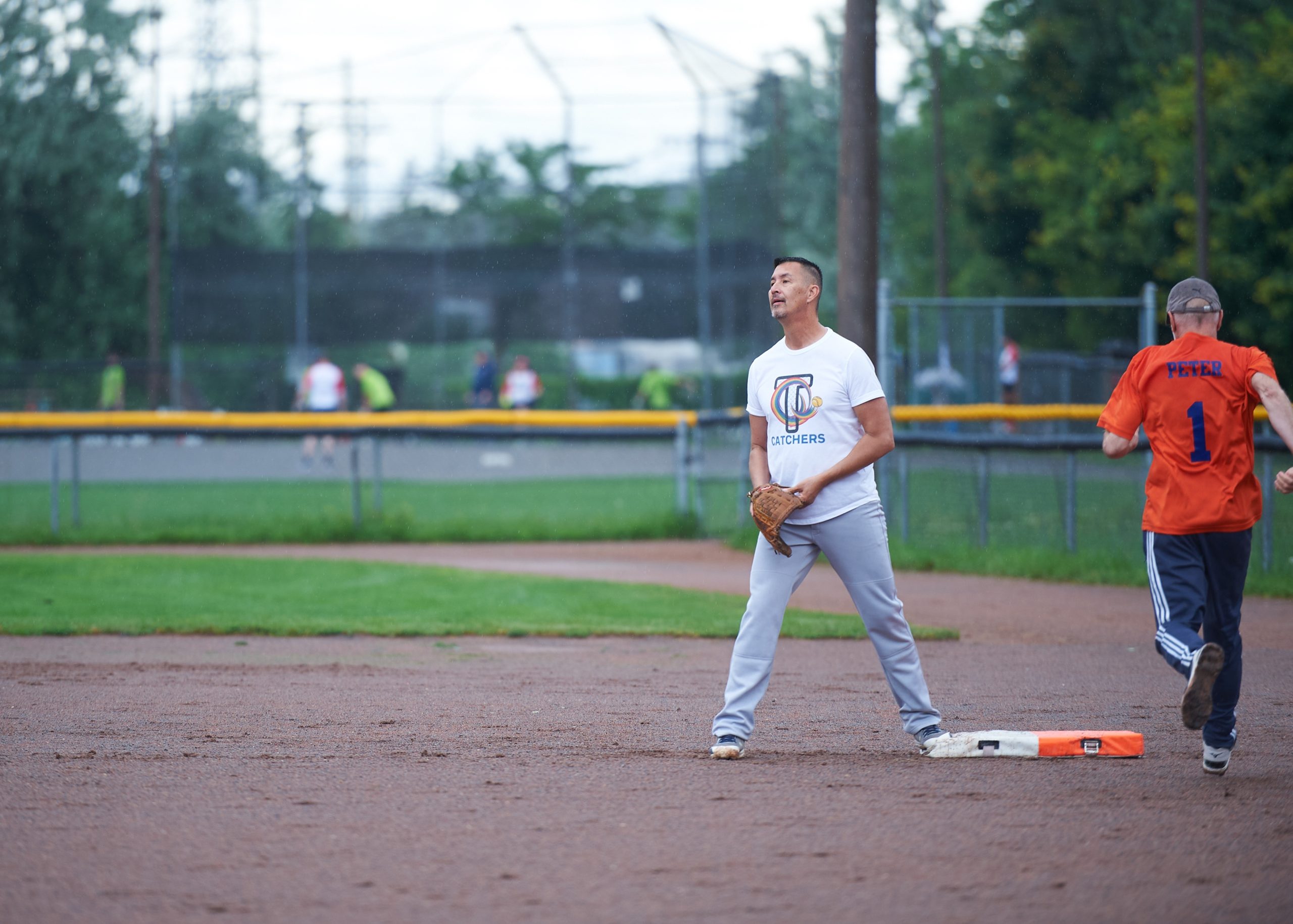 A man wearing a white shirt with the word ‘catchers’ on it waits for the ball. 