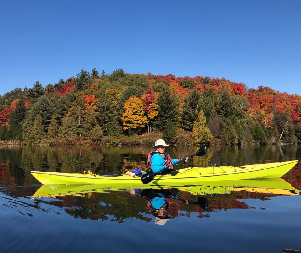 Linda in a Like green kayak paddles in a lake. Threes with fall colours are in the background on a sunny day.