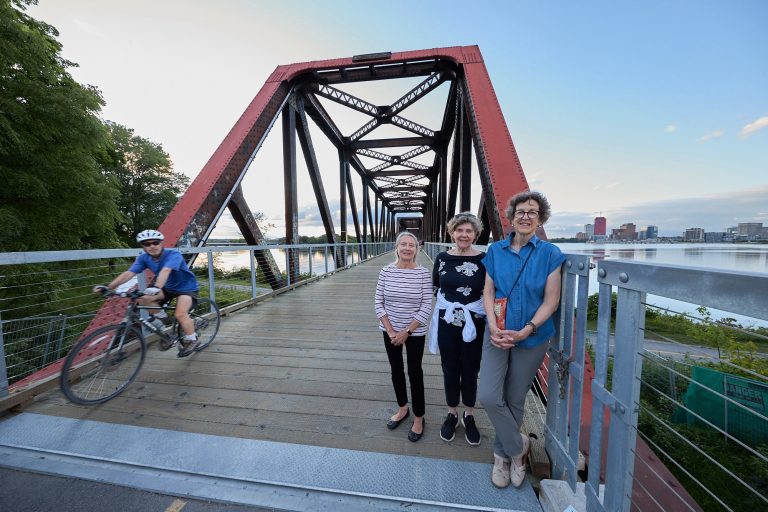 Three women pose for a photo on the Chief William Commanda Bridge. A person on a bike passes by.