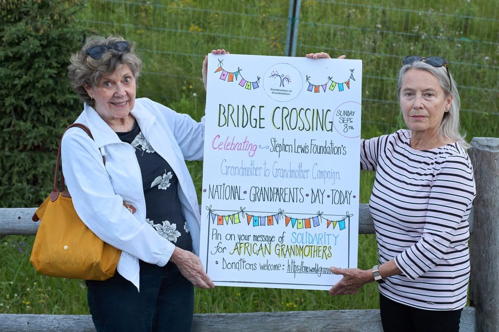 Two women hold a sign next to a wooden fence. The sign is promoting the bridge crossing.