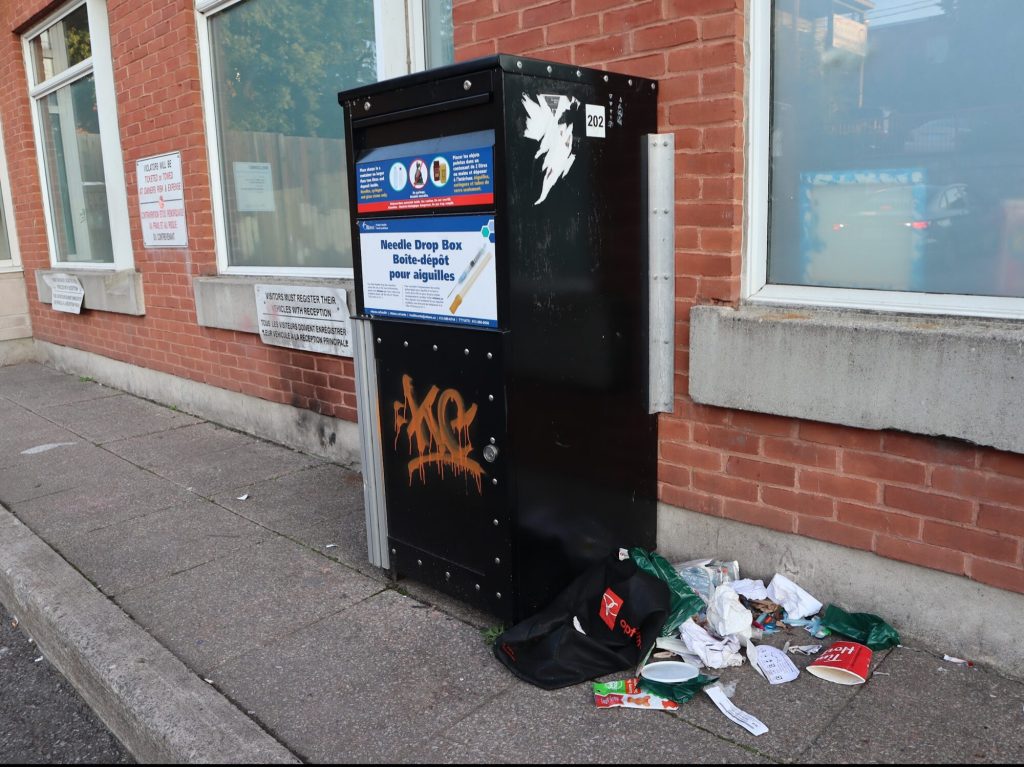 A needle drop box with garbage littered around it.