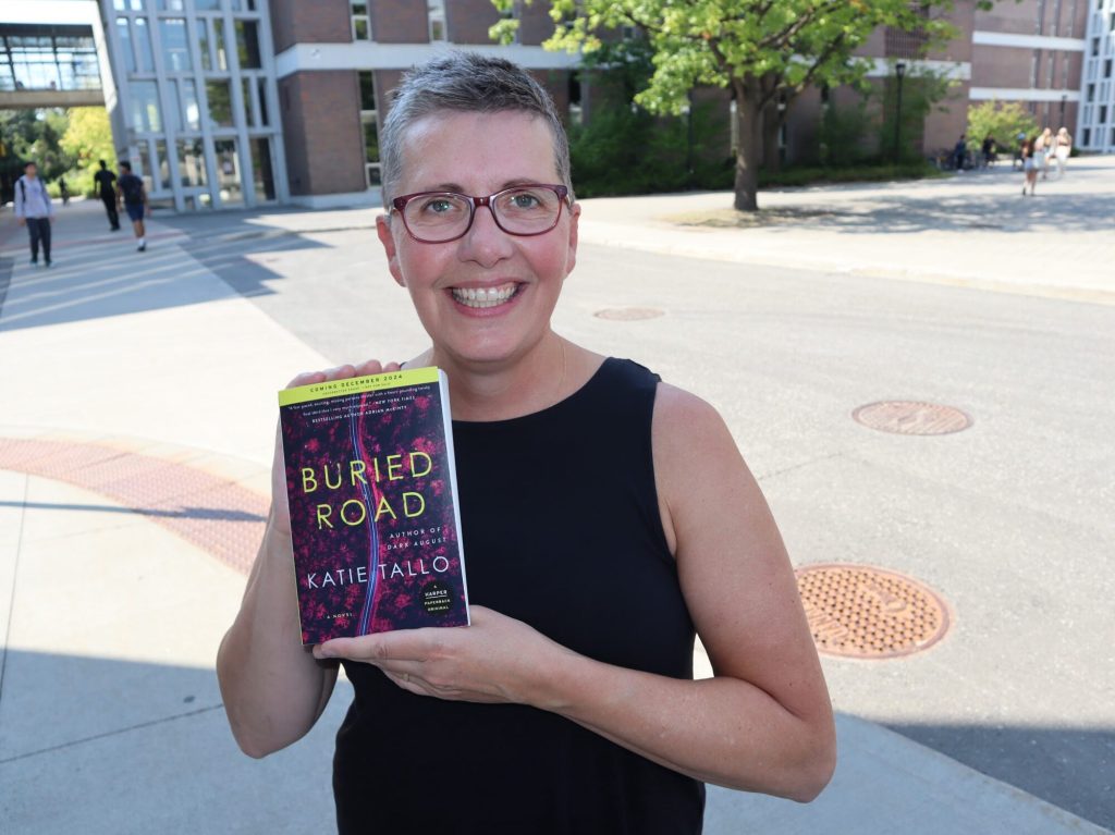 Katie stands outside of buildings at Carleton University and holds up a copy of her book.