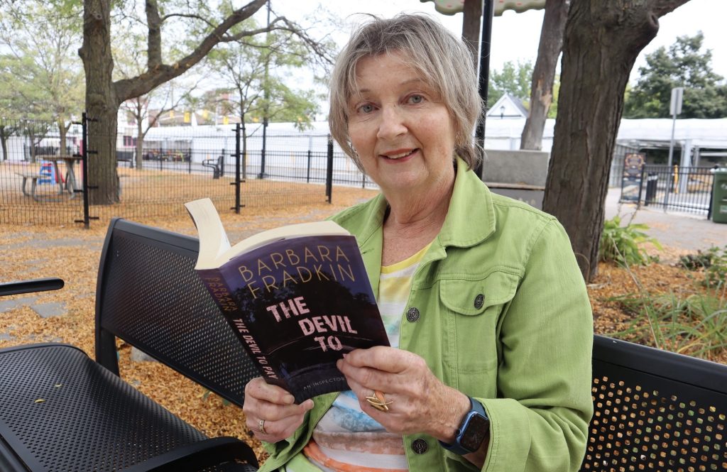 Barbara looks at the camera and smiles as she sits on a bench and reads a book.