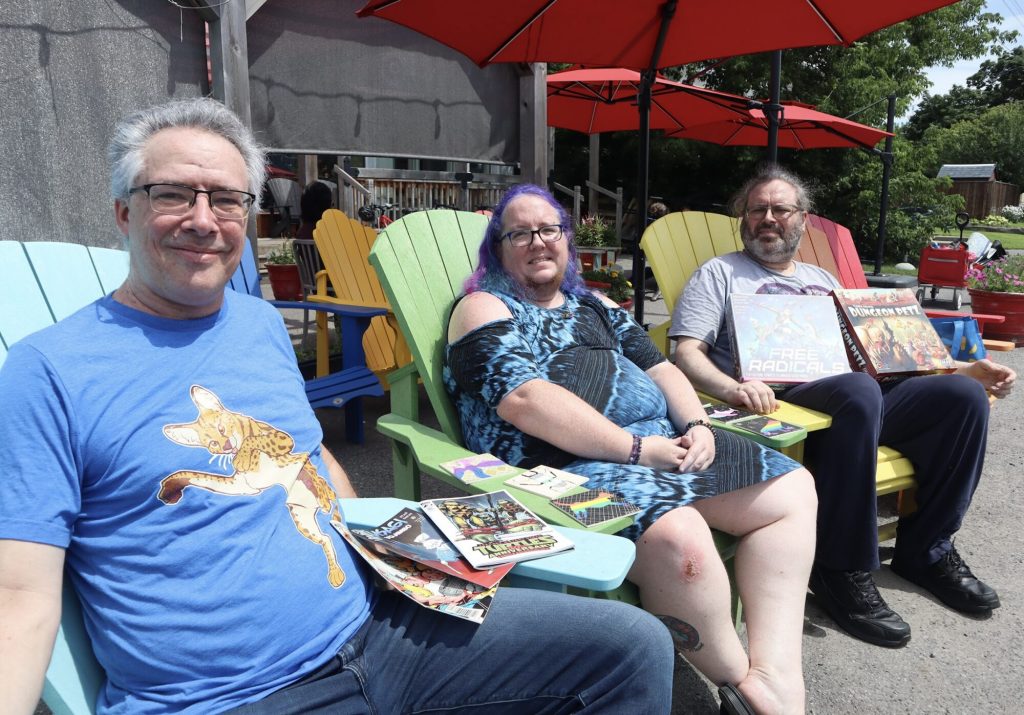 Three people pose for photos in colourful Mishima chairs. They are holding board games and other items.