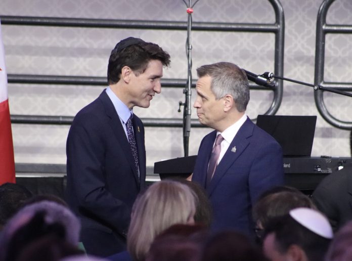 Prime Minister Justin Trudeau and Ottawa Mayor Mark Sutcliffe stand close as they engage in conversation. A crowd of people are seen to the front of them.