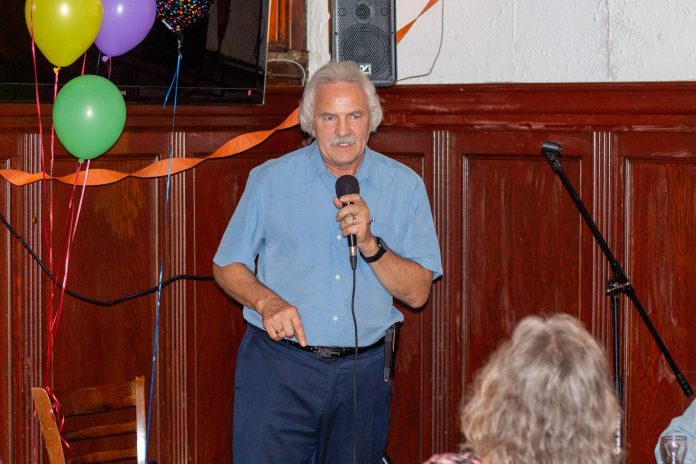 John who’s wearing a blue shirt holds a microphone and speaks to a crowd which is not pictured.