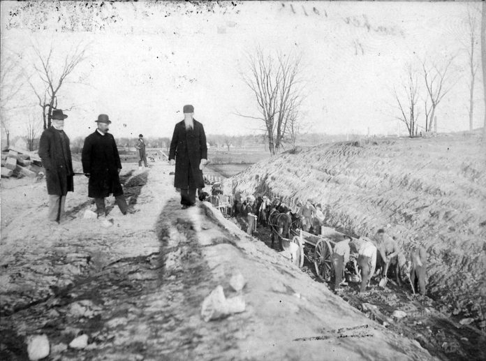 A black and white photo of three men standing next to a ditch.