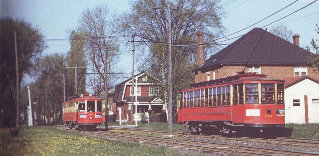 A coloured photograph of two red streetcars passing through a residential area.